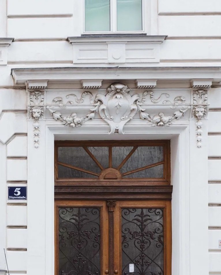Historisches Portal eines Altbaus mit verzierten Holztüren und einem dekorativen Giebelrelief aus weißem Stuck an einer hellen Fassade.