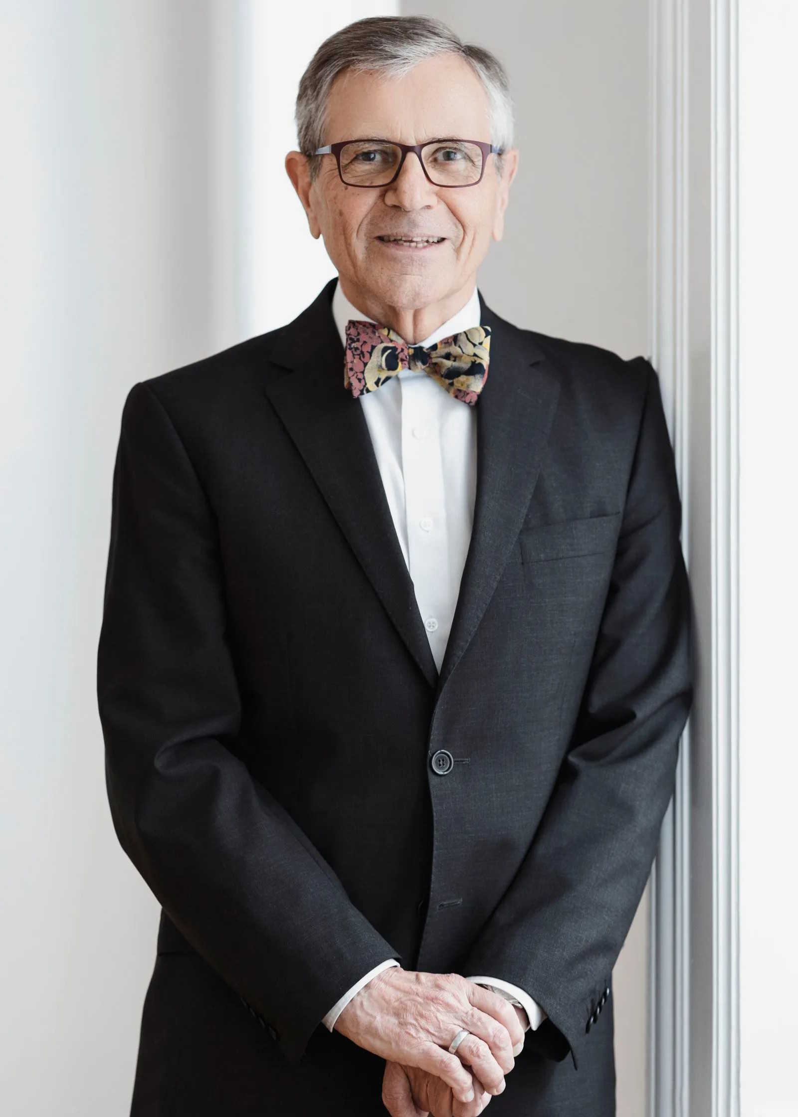 Porträt eines freundlich lächelnden Mannes in einem dunklen Anzug mit einer gemusterten Fliege und Brille.