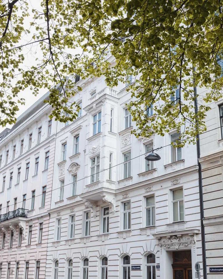 Klassisches helles mehrstöckiges Zinshaus mit historischer Fassade, Balkonen und hohen Fenstern, eingerahmt von grünen Bäumen