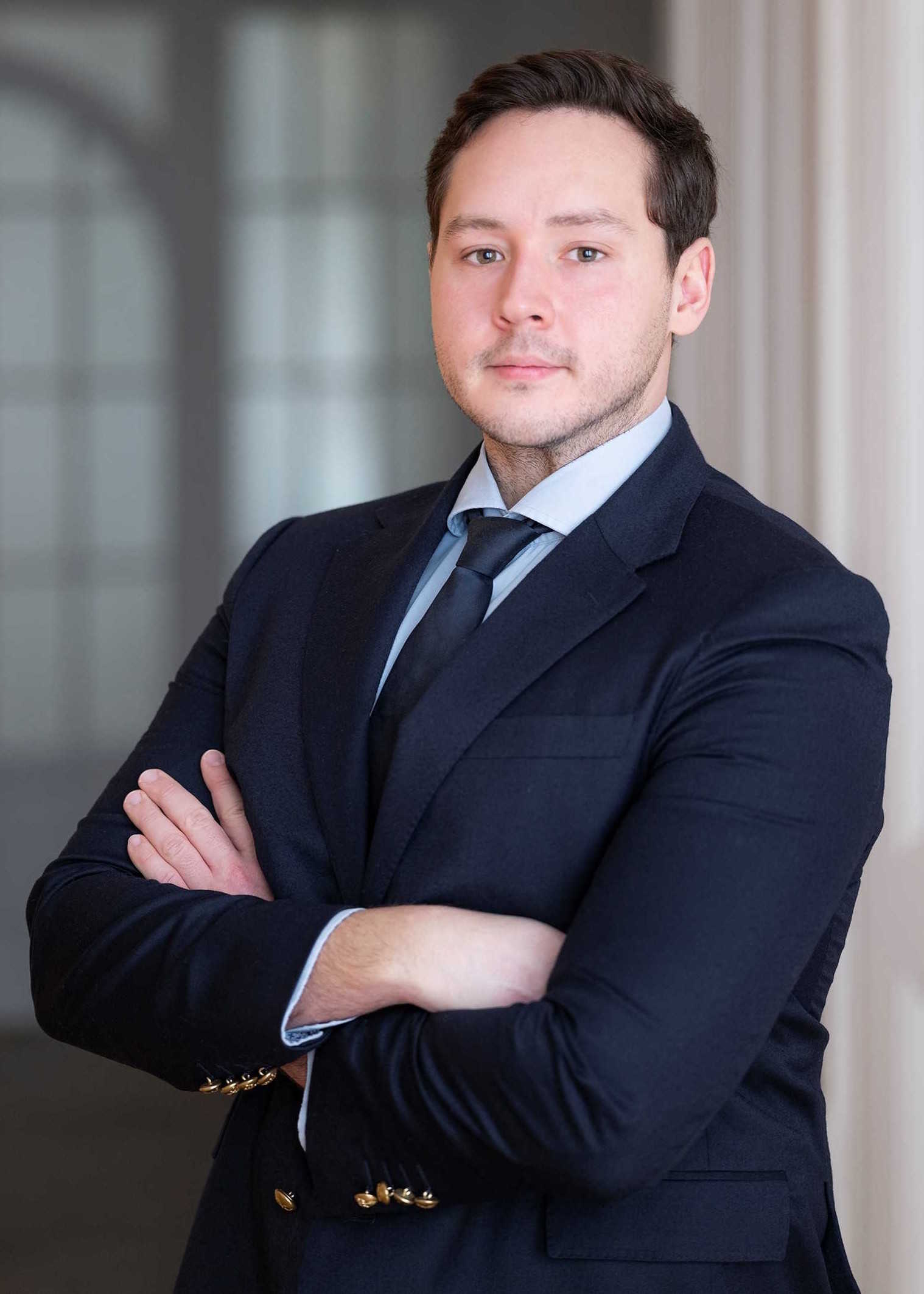 Porträt eines jungen Mannes in einem dunkelblauen Sakko mit Goldknöpfen und Krawatte, der mit verschränkten Armen professionell in die Kamera blickt.